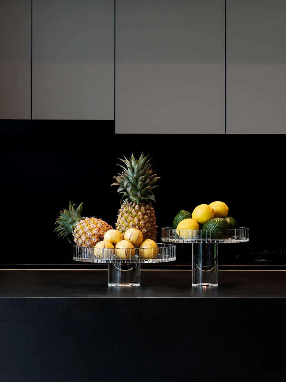 Minimalist home kitchen layout shot of two SHIJI M crystal fruit plates with fresh fruits against grey cabinet and dark countertop