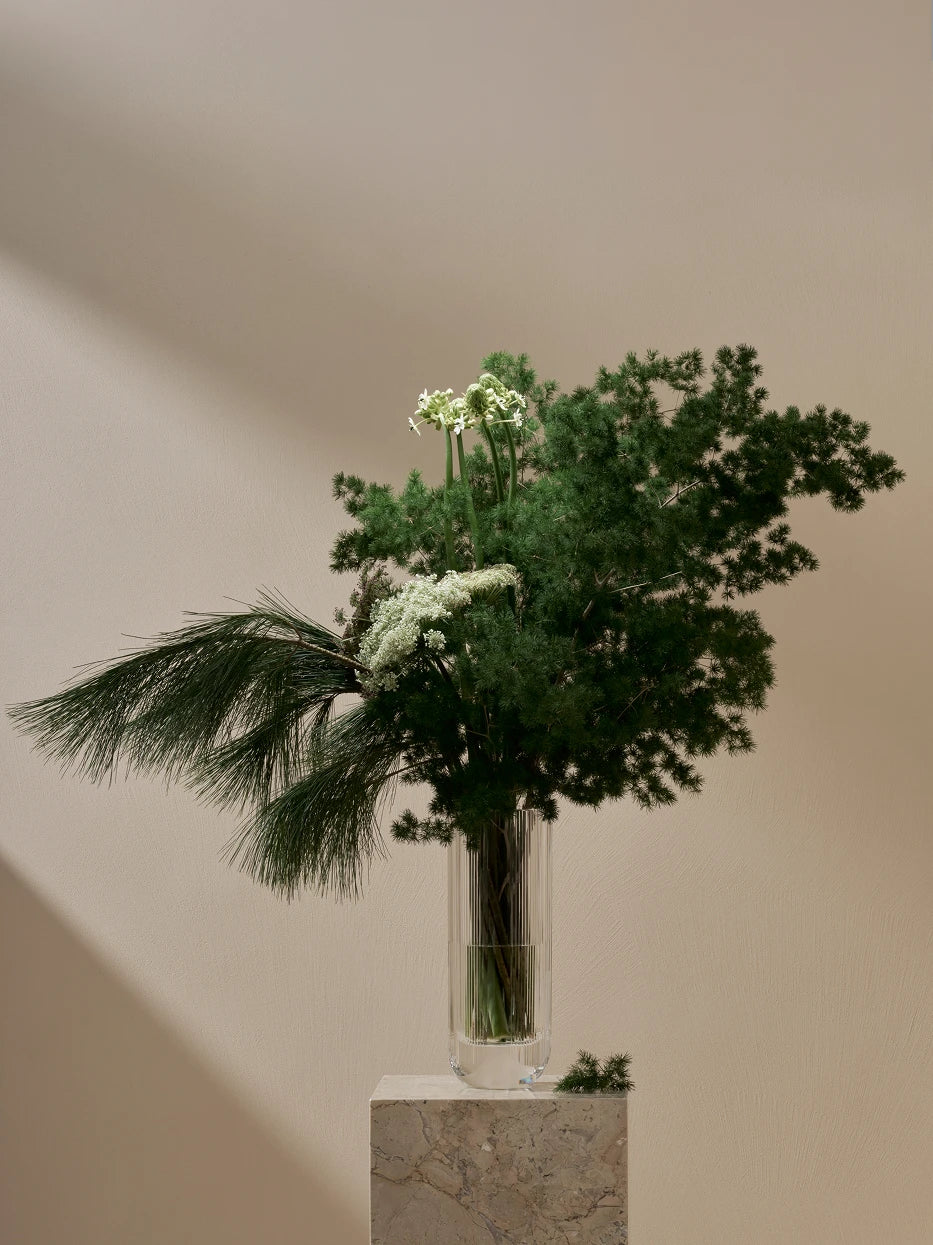 Minimal artistic furnishing shot of SKYSCRAPER crystal vase with dense pine foliage & tiny white blooms on travertine stone plinth