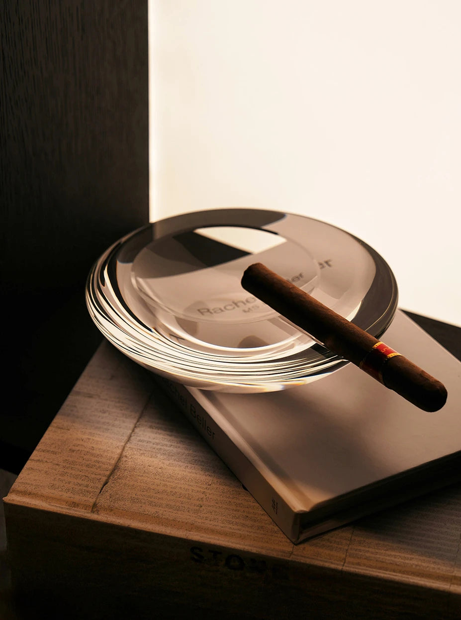 Warm tone still life close-up of large WATERDROP L ashtray with cigar on warm wood side table, soft side light showing multi-layer crystal refraction