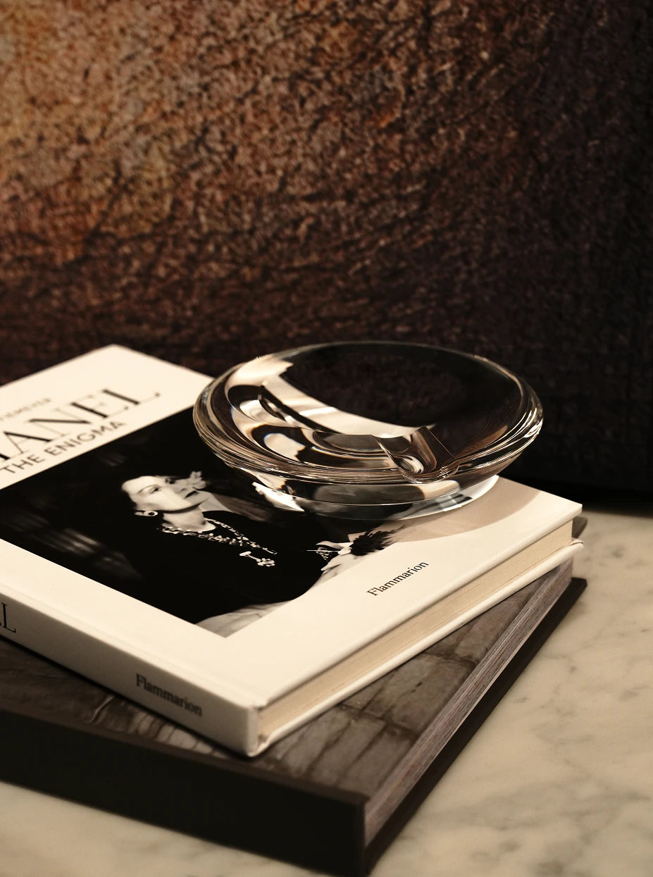 Vintage still life close-up of WATERDROP M crystal ashtray stacked on black-and-white fashion art books against textured dark backdrop
