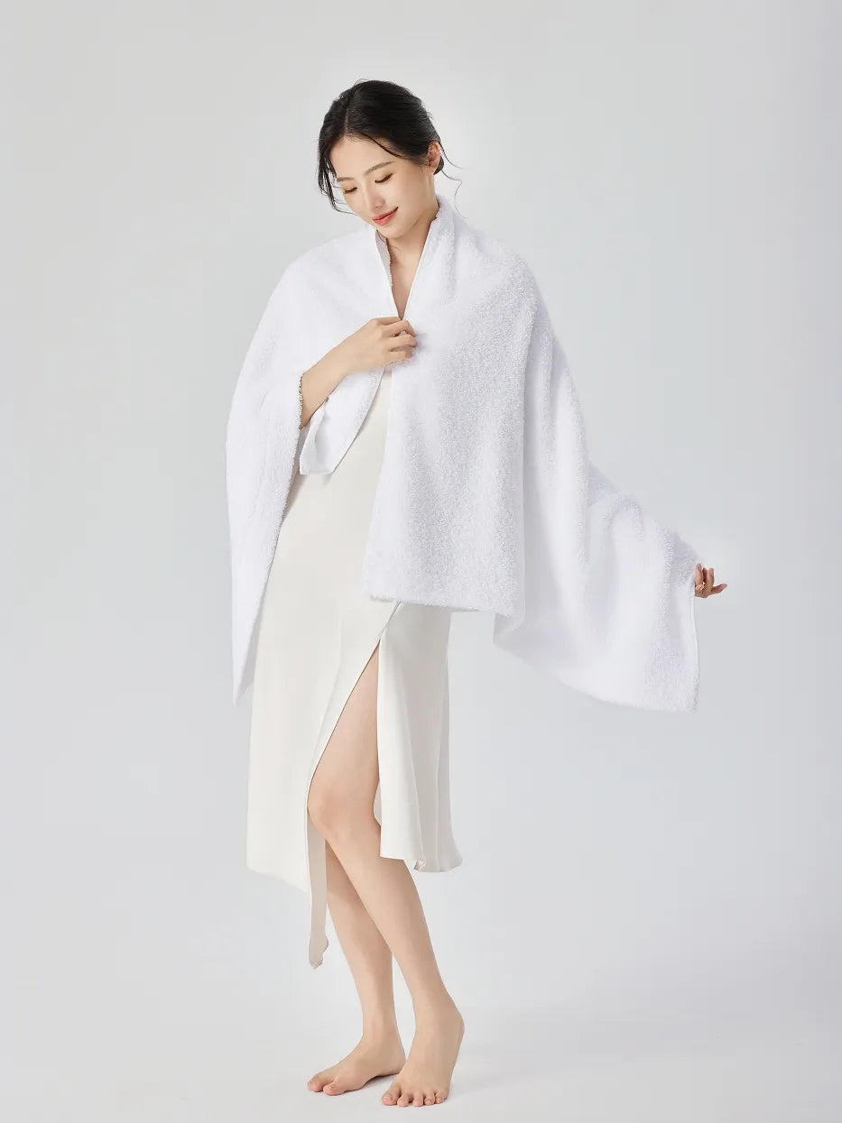 Barefoot woman standing, draping RESONG white cotton terry bath towel over shoulders like a shawl, looking down softly, white slip dress with side slit, white studio background

