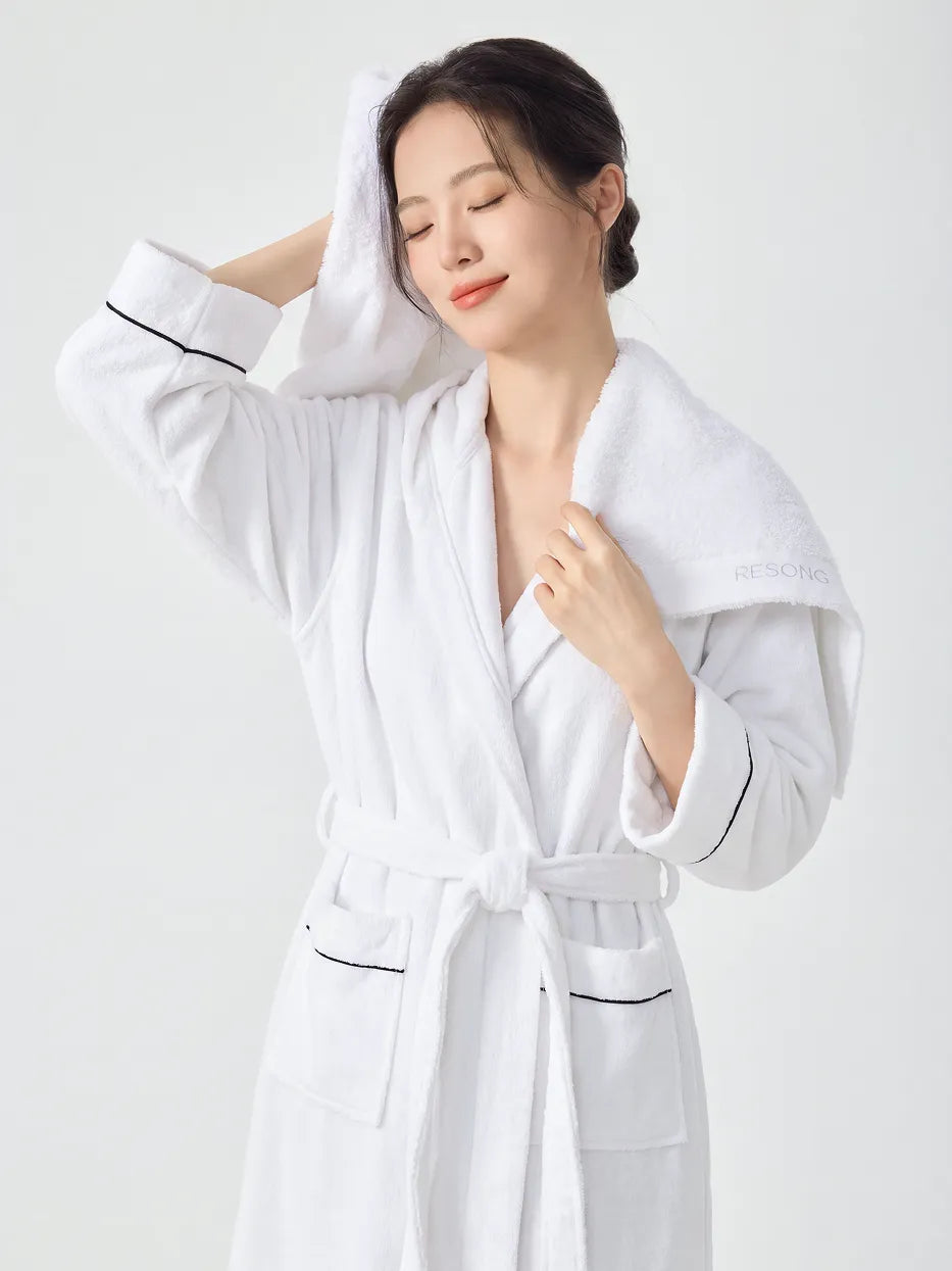 Woman in white hooded bathrobe with black piping, eyes closed drying hair with a RESONG white cotton terry face towel draped over shoulder, embroidered brand label visible, soft fluffy face towel gift set, white studio background

