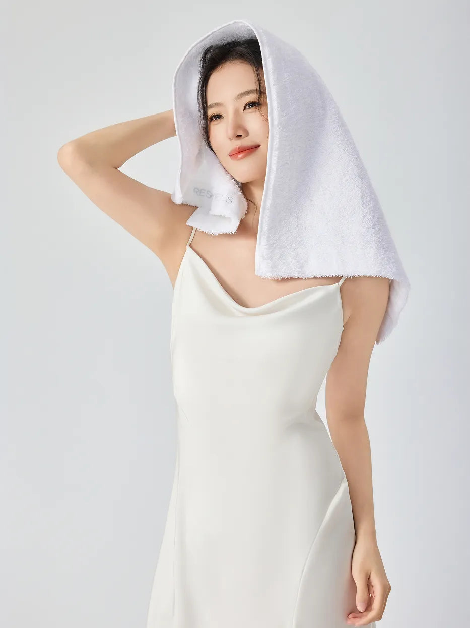 Woman in white slip dress draping RESONG white cotton terry face towel over head like a hood, showing inner woven brand label, soft fluffy face towel gift set, white studio background
