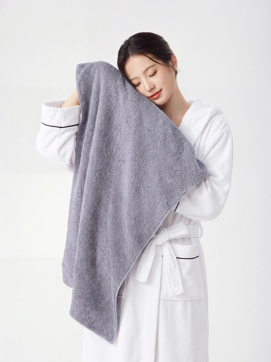 Woman wearing RESONG white hooded terry cloth bathrobe with black piping detail, eyes closed gently pressing a grey cotton bath towel against her cheek, luxury bath towel and bathrobe gift set, white studio background
