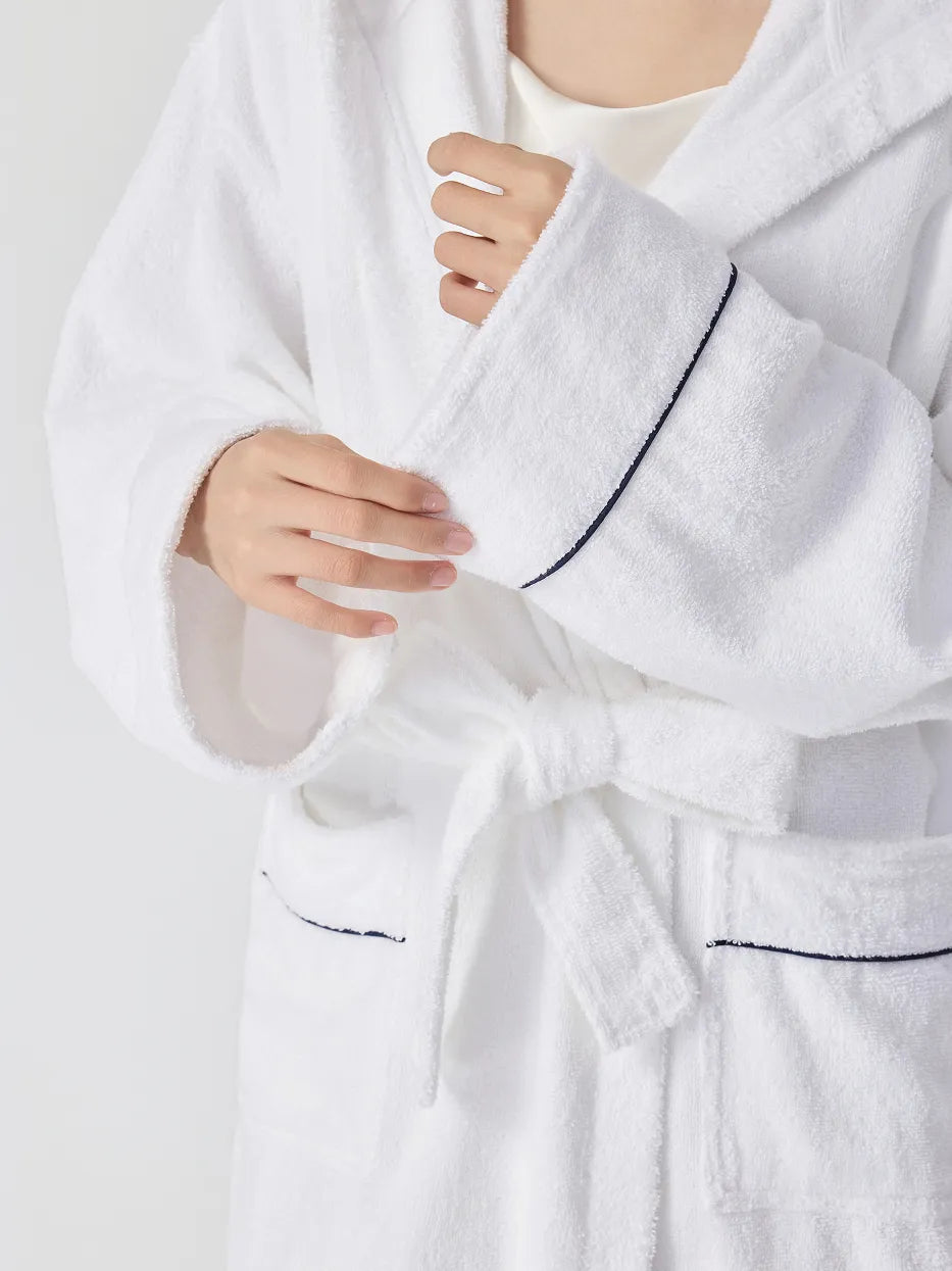 Close-up of RESONG white hooded terry cloth bathrobe showing navy piping on cuff and pockets, matching belt tie, hands adjusting sleeve, soft absorbent cotton texture, white background

