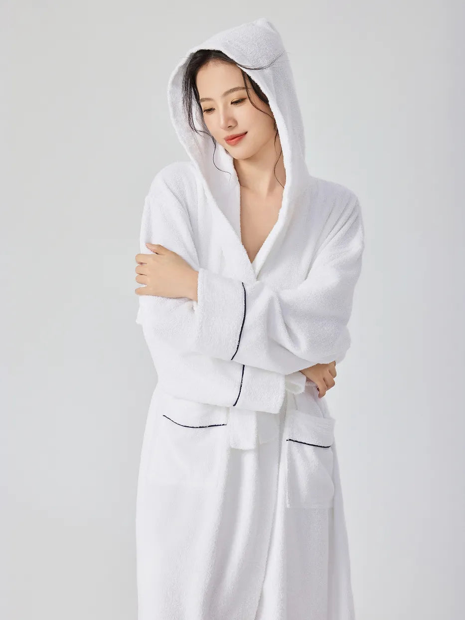 Woman wearing RESONG white hooded terry cloth bathrobe with hood up, half body, arms crossed, smiling softly and looking down, navy piping on cuffs and pockets, soft absorbent cotton, white studio background
