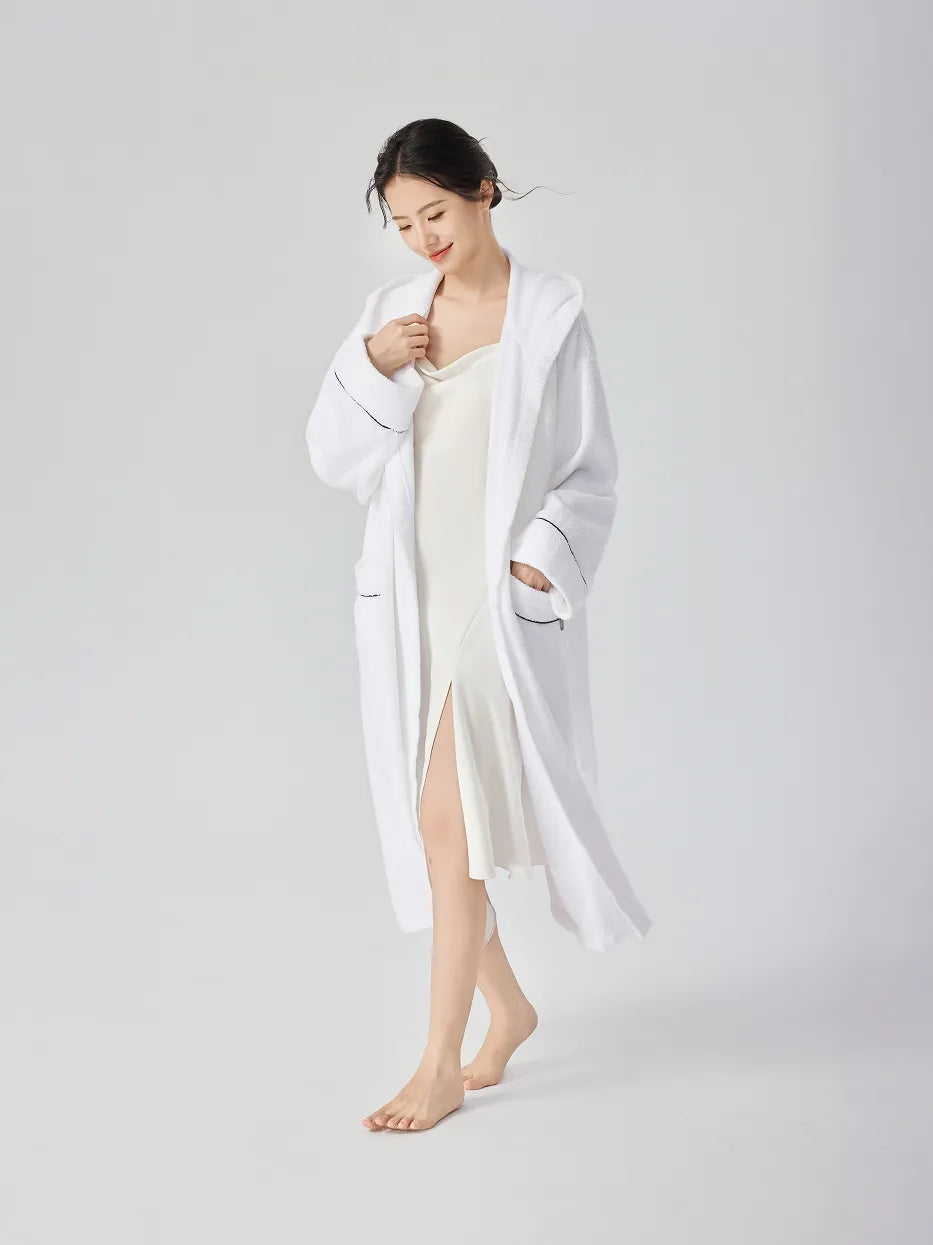 Barefoot woman walking in RESONG white hooded terry cloth bathrobe worn open over white slip dress, one hand in pocket, navy piping on cuffs, relaxed long fit, white studio background

